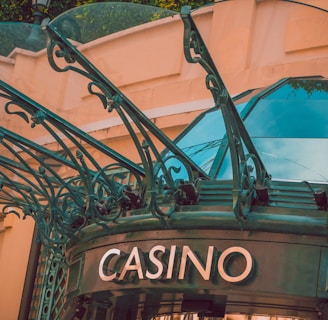 The image displays the entrance to a casino, characterized by detailed and ornate architectural elements with green metal structures and a sign that reads 'CASINO'. The background includes a glass dome and a peach-colored wall.