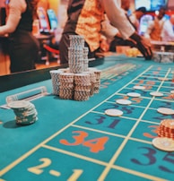 A casino scene with a roulette table features prominently, covered in green felt with colorful numbers and betting squares. Several stacks of poker chips are placed on different numbers, and the hands of several people are visible as they interact with the table. In the background, there are slot machines and more people, creating a vibrant and busy atmosphere.