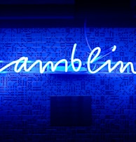 A bright blue neon sign spells out the word 'gambling' in cursive script. The background features a textured wall covered in domino tiles, also illuminated by the blue light, creating a vivid and visually striking scene.