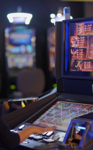 A person is seated at a slot machine console in a casino, focusing intently on the game displayed on the screen. The background shows several other illuminated gaming machines, adding a vibrant and colorful atmosphere to the scene.