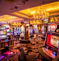 A vibrant casino interior filled with rows of colorful slot machines, each illuminated with bright lights and displays. The ceiling is adorned with elegant chandeliers and golden drapes, creating a luxurious atmosphere. The patterned carpet adds to the elaborate design of the space.