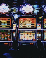 A row of colorful slot machines lit with bright neon lights, showcasing various graphical elements on the screens, indicating different games and jackpots. The atmosphere suggests a lively casino setting, bustling with excitement.