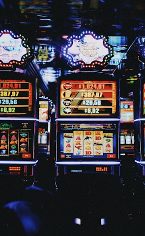 A row of colorful slot machines lit with bright neon lights, showcasing various graphical elements on the screens, indicating different games and jackpots. The atmosphere suggests a lively casino setting, bustling with excitement.