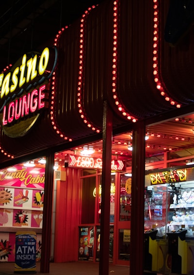 A brightly lit casino lounge entrance adorned with neon lights. The exterior features colorful signage including the words 'Casino' and 'Lounge' in vivid colors, with additional lights framing the building. Inside, a glimpse of a claw machine filled with plush toys and arcade games is visible. The area exudes an inviting yet bustling atmosphere typical of entertainment venues.
