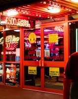 A brightly lit entrance to a casino with neon signs, including one advertising a £500 jackpot. The doors have clear signage indicating entry is restricted to those over 18. A person in a red jacket stands to the right, possibly interacting with a machine or display.