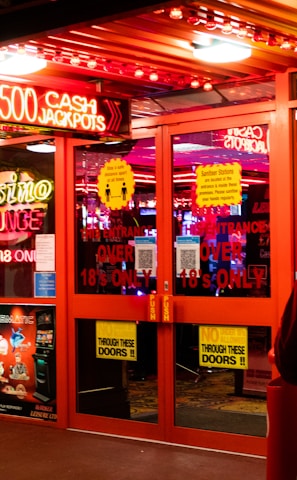 A brightly lit entrance to a casino with neon signs, including one advertising a £500 jackpot. The doors have clear signage indicating entry is restricted to those over 18. A person in a red jacket stands to the right, possibly interacting with a machine or display.
