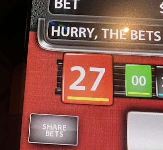 A digital display screen showing a casino betting interface, with text prompting to hurry bets and a highlighted number 27 in red. There are options for betting amounts and a 'share bets' button.