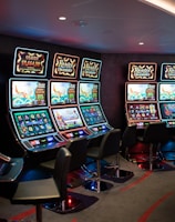 A row of brightly lit slot machines in a casino setting, each with colorful displays and various game themes. The machines are lined up against a dark wall and are unoccupied, with cushioned chairs placed in front of them. The area is dimly lit except for the glow from the screens, creating a vibrant and engaging atmosphere.