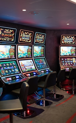 A row of brightly lit slot machines in a casino setting, each with colorful displays and various game themes. The machines are lined up against a dark wall and are unoccupied, with cushioned chairs placed in front of them. The area is dimly lit except for the glow from the screens, creating a vibrant and engaging atmosphere.