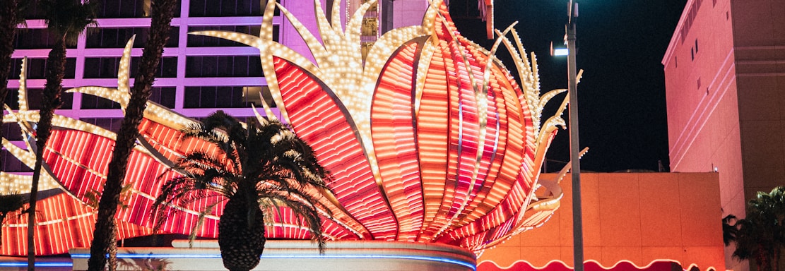 A brightly lit casino with neon lights in vibrant red, pink, and blue, featuring a large flamingo signage. Palm trees are visible in the foreground with vehicles passing by on the street at night.