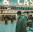 An older man with a gray beard and wearing a suit and hat stands in front of a brightly lit casino. The background features a large illuminated sign reading 'CASINO' and 'SLOTS' among other words, with numerous lights outlining the entrance. Several other people are visible in the background, engaged in various activities, some walking, some standing, and one looking at something on display.