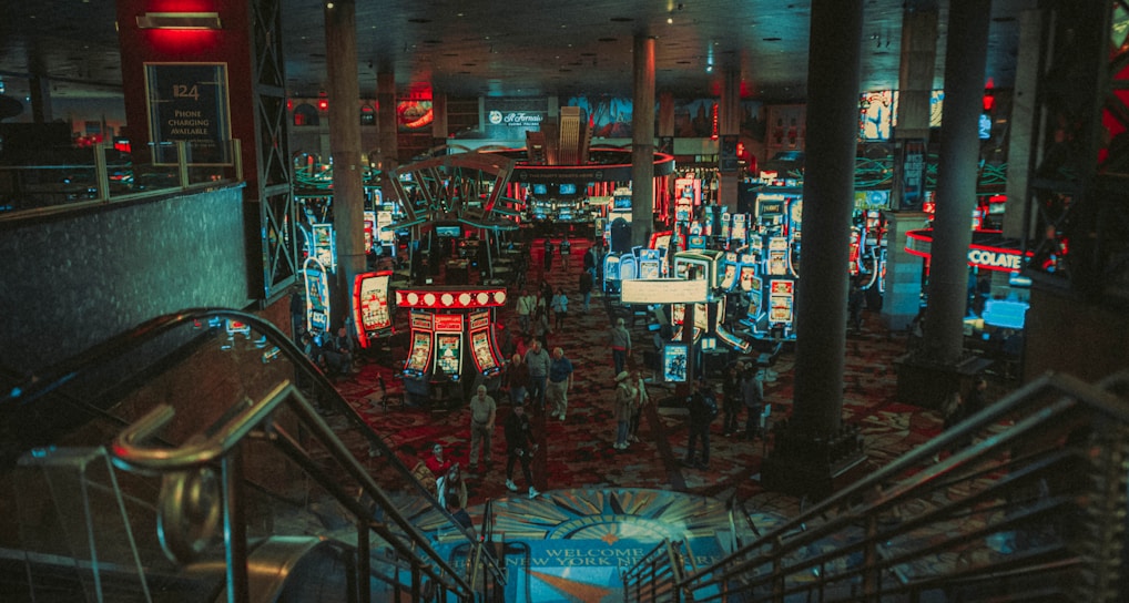 A bustling casino floor filled with groups of slot machines and people walking around. The atmosphere is vibrant with neon lights and illuminated signs. The interior has a ceiling supported by large pillars and there are stairs leading down to the gaming area.